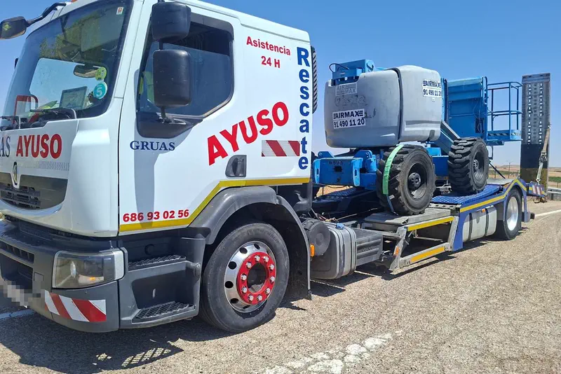 Grúa de plataforma de Grúas Ayuso, rotulada Rescate y Asistencia 24 H, cargando una plataforma elevadora en la carretera.