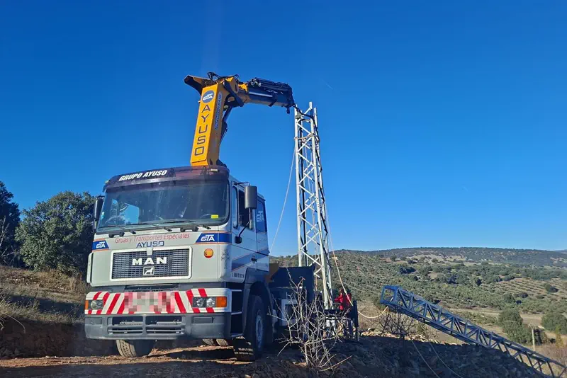 Camión grúa con pluma articulada amarilla rotulada Ayuso izando un tramo de torreta eléctrica.