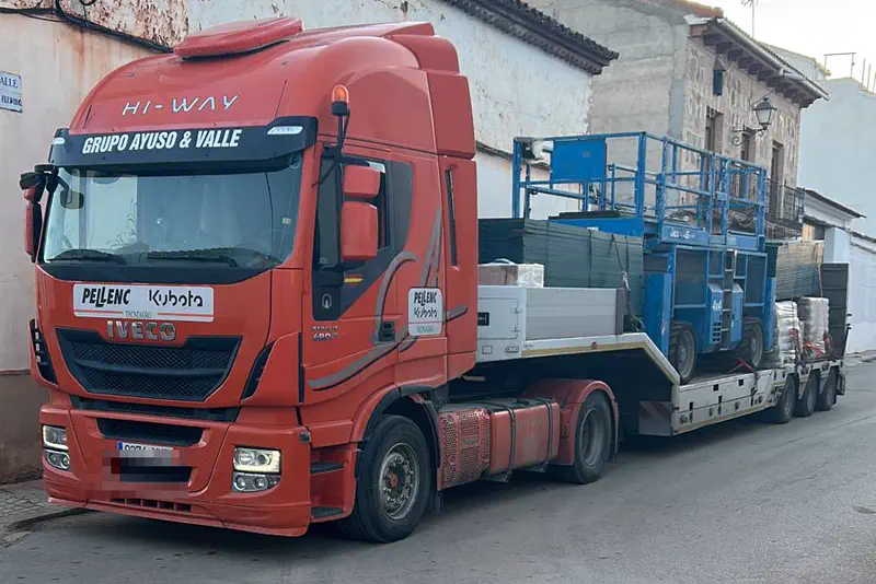 Tractora con góndola cargada con una plataforma elevadora y material paletizado en una calle de pueblo.